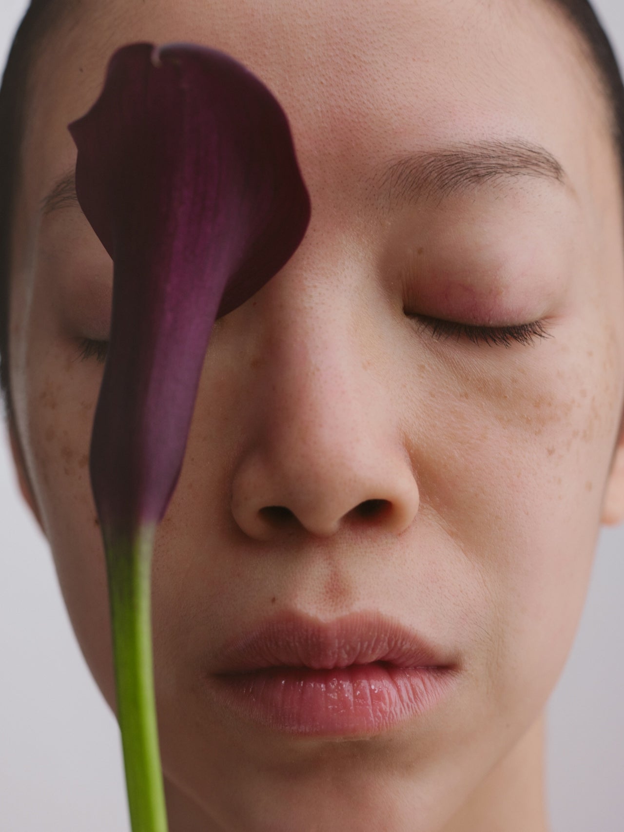 Close-up of a person with a purple flower covering one eye against a neutral background