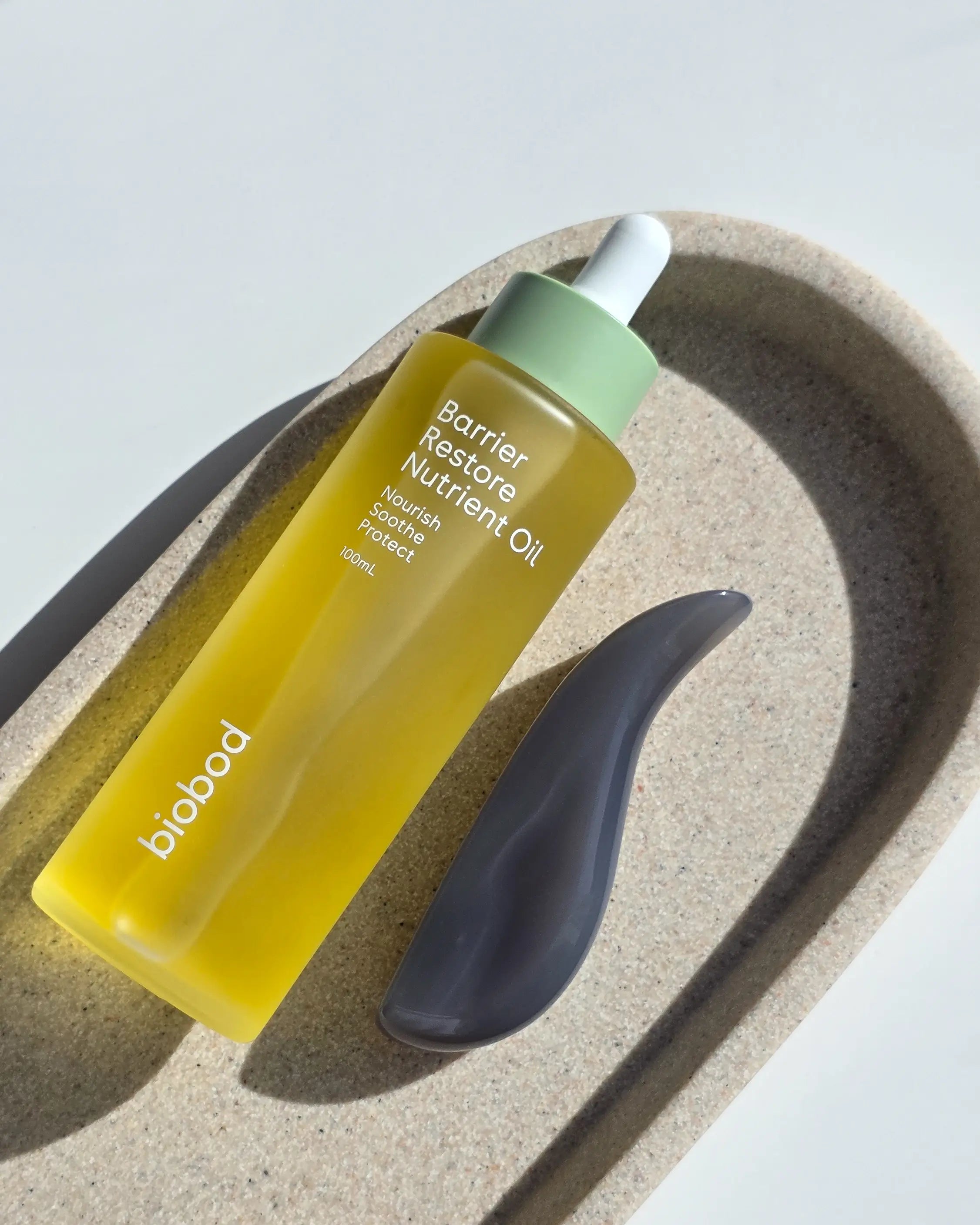 Yellow bottle of Barrier Restore Nutrient Oil by biobod with gua sha with sensitive skin on a textured beige surface with a white background