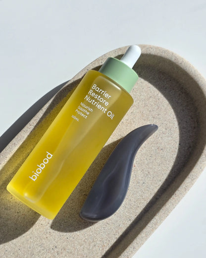 Yellow bottle of Barrier Restore Nutrient Oil by biobod with gua sha with sensitive skin on a textured beige surface with a white background